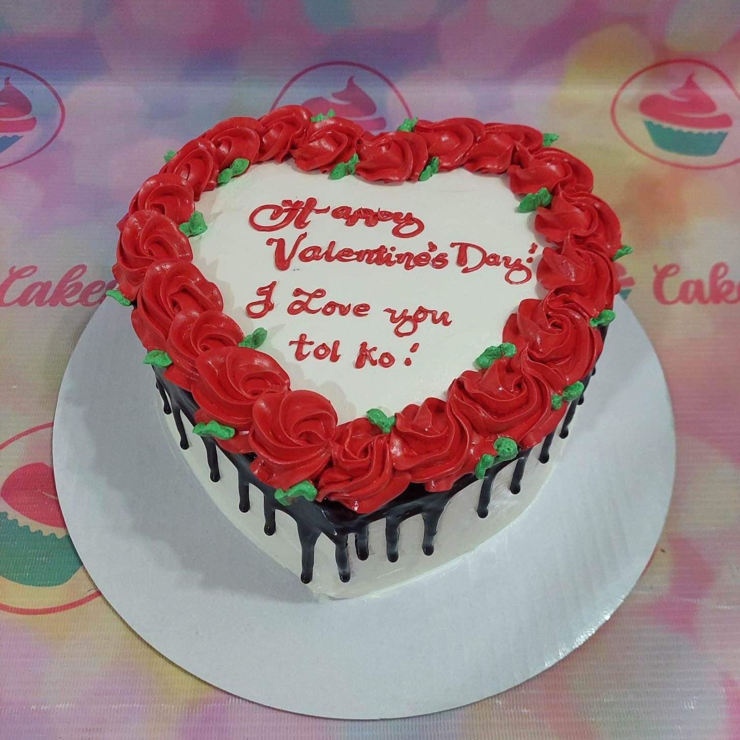 Heart-shaped white cake with red icing roses, chocolate drip, and a Valentine's Day message.