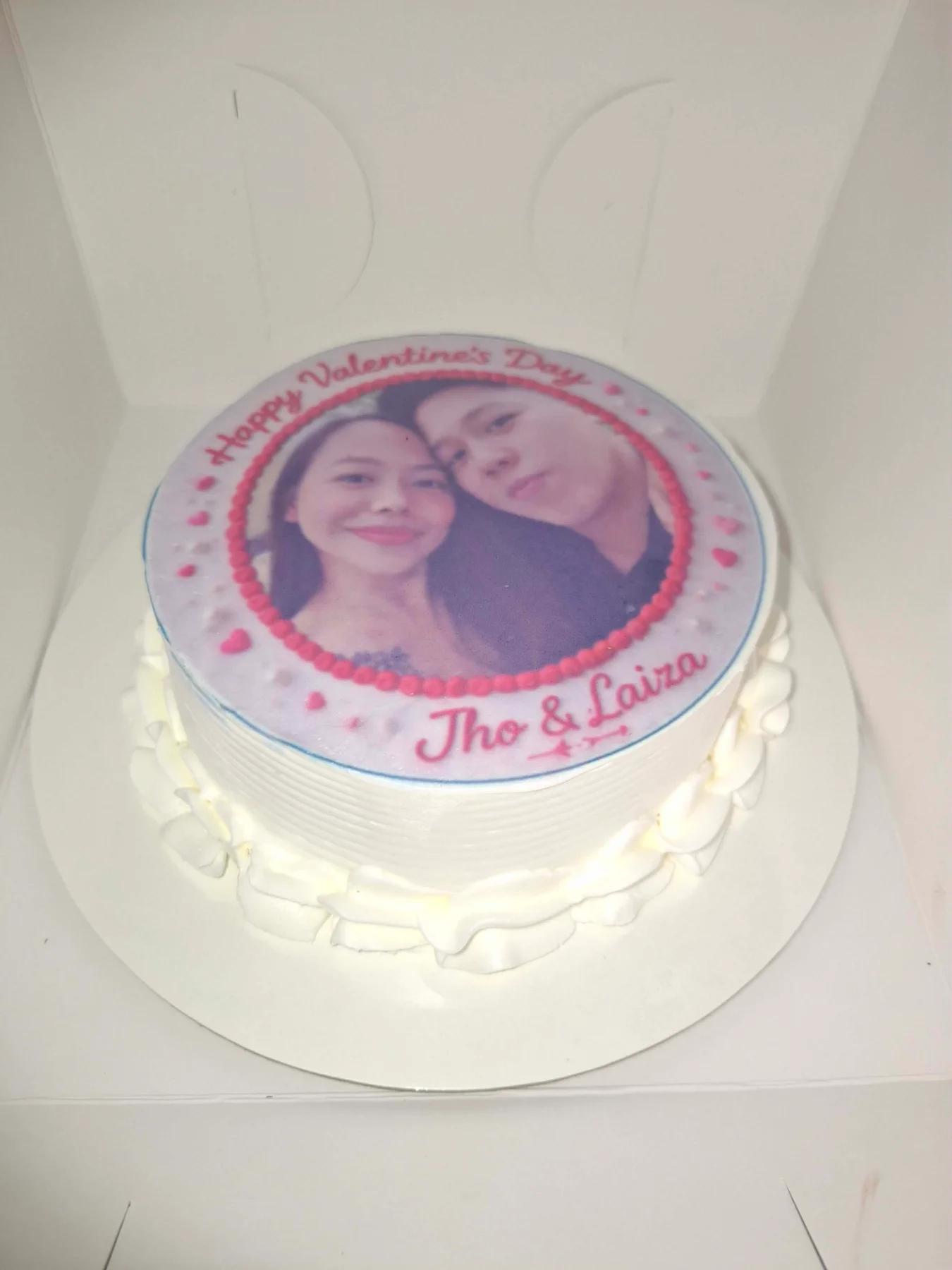 White round cake with a large edible photo print of a couple, pink icing borders, and a Happy Valentine's Day message.