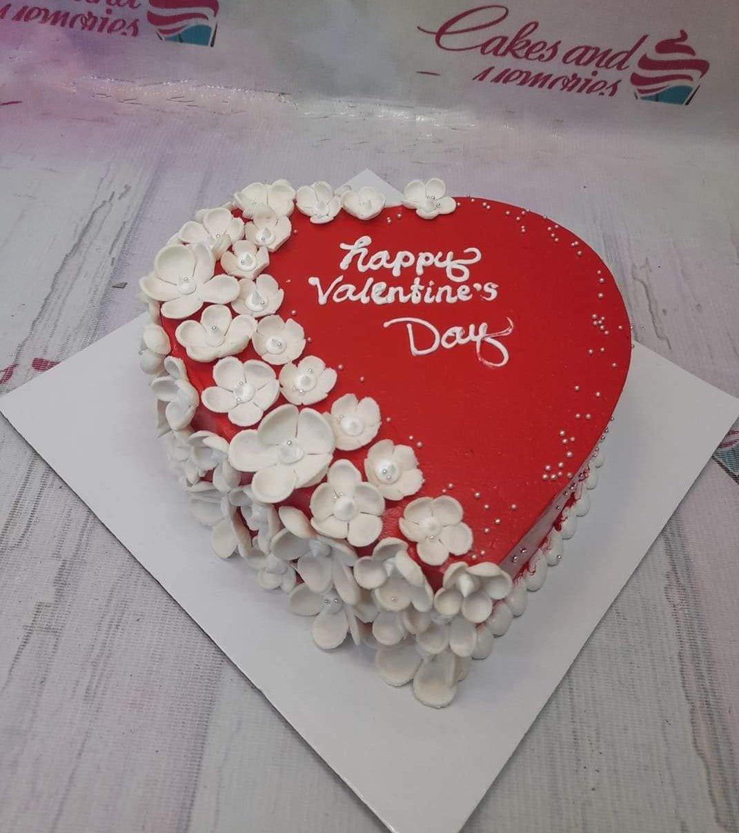 Heart-shaped red cake with white gumpaste flowers and Happy Valentine's Day message in white icing.