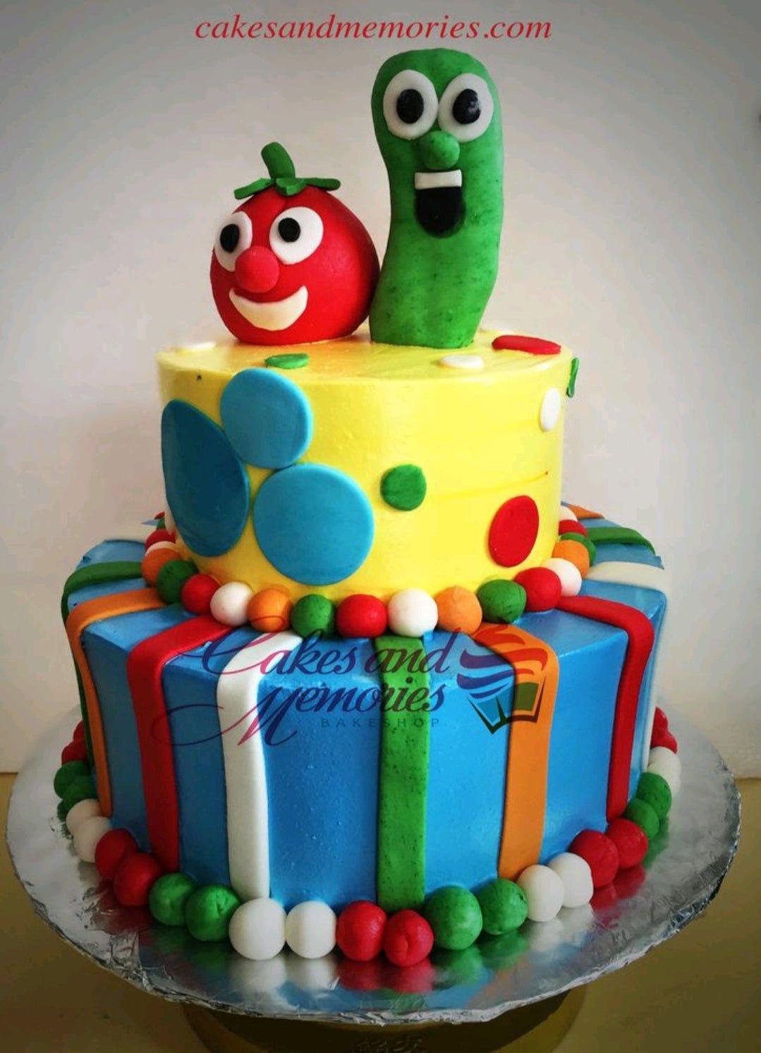 Two-tier VeggieTales themed fondant cake with Bob the Tomato and Larry the Cucumber toppers on yellow and blue tiers with colorful stripes.