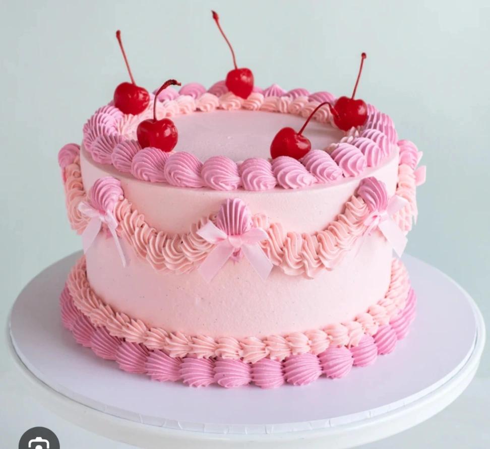 Vintage-style pink cake with intricate icing ruffles, pink bows, and fresh red cherries on top.