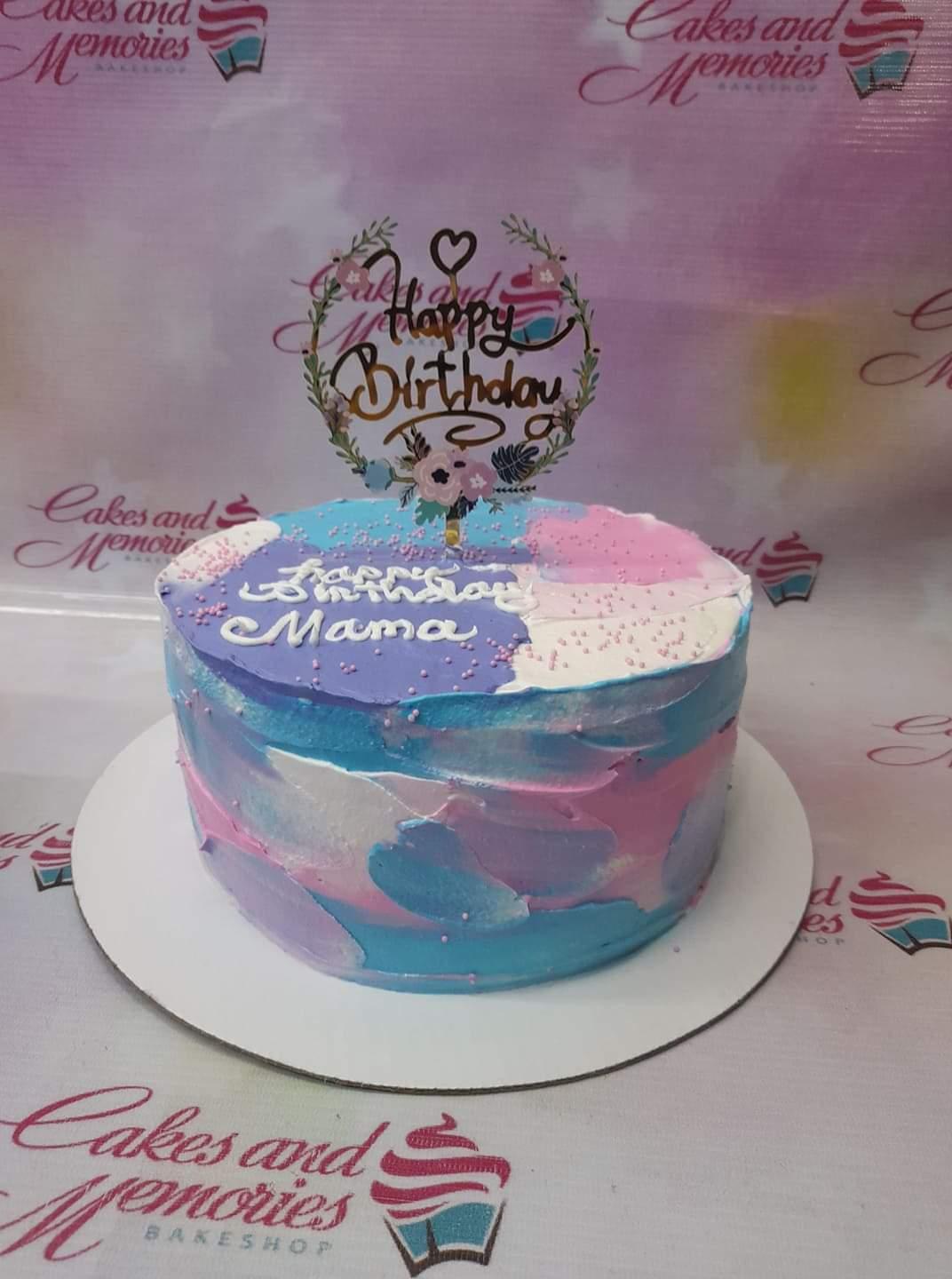 A single-tier birthday cake with a watercolor-style palette knife icing in blue, pink, purple, and white, topped with a floral Happy Birthday topper.