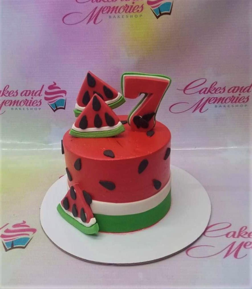 Red watermelon themed birthday cake with green and white base stripes, black seed accents, and fondant watermelon slice toppers.