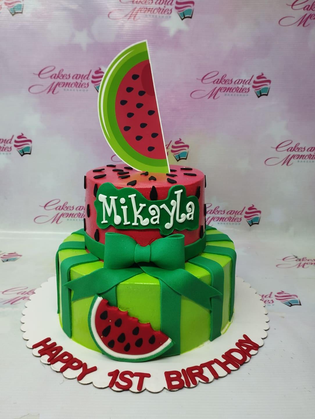 Two-tier watermelon themed birthday cake with green striped bottom tier and red top tier featuring black seeds and a large watermelon slice topper.