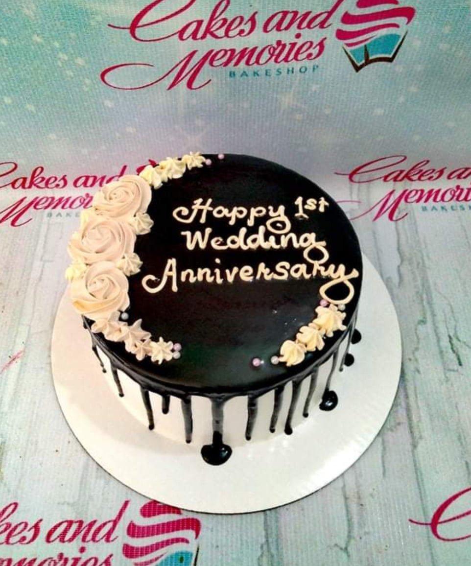 Single tier white cake with black chocolate drip, white icing roses, and a Happy 1st Wedding Anniversary message.