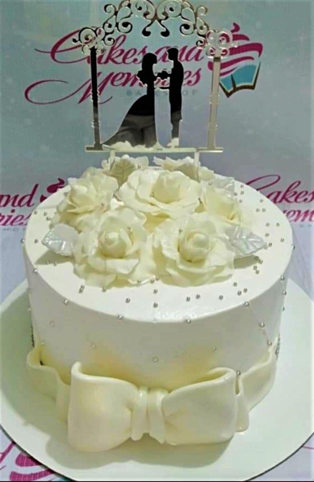 White 1-tier wedding or anniversary cake with large white gumpaste roses, silver dragees, a large white bow, and a silver couple topper.
