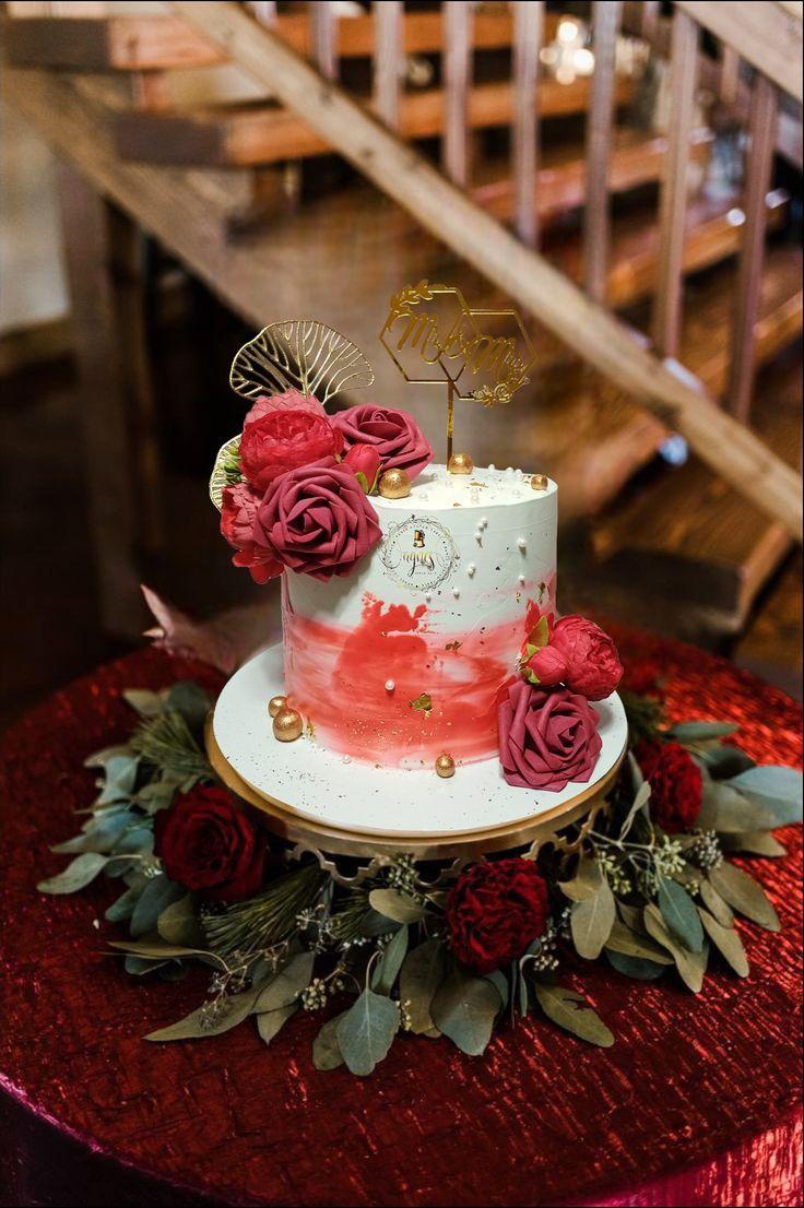 Elegant white cake with red watercolor icing, gold leaf accents, and deep red artificial roses topped with a gold Mr & Mrs topper.