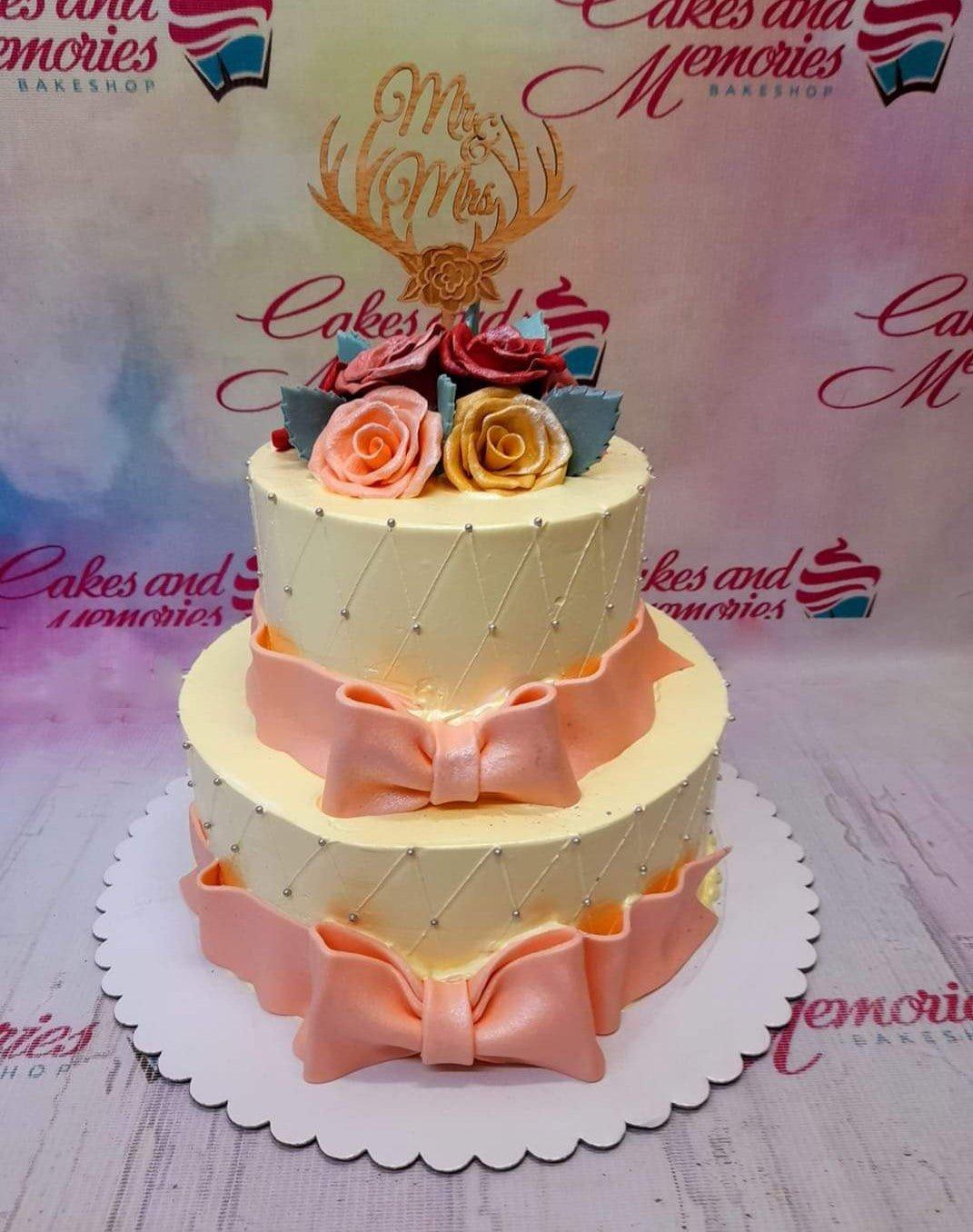 Elegant two-tier ivory wedding cake with peach fondant bows, silver dragees, and a wooden Mr & Mrs topper with edible roses.