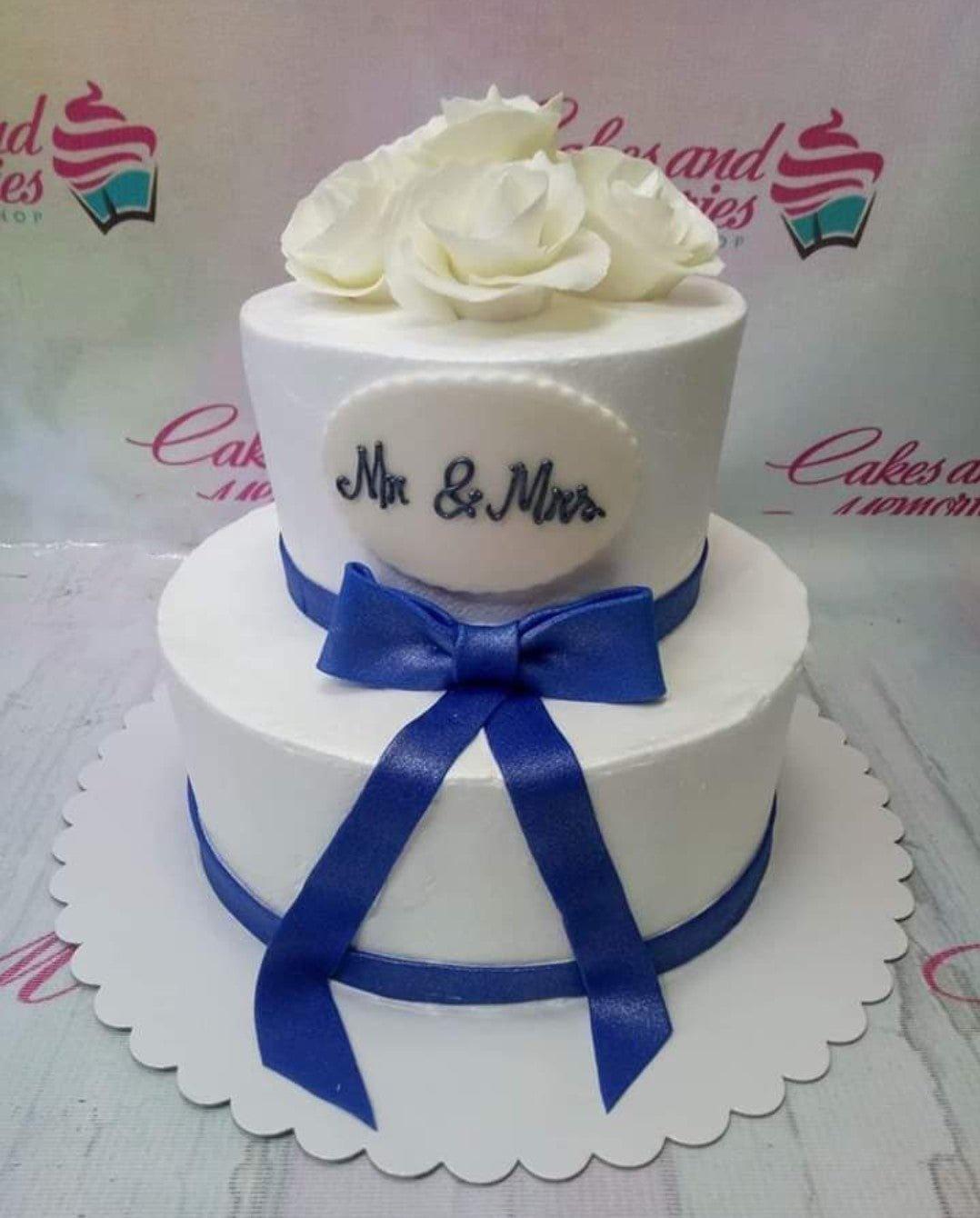 Elegant white 2-tier wedding cake with white edible roses and a large royal blue satin-style bow.