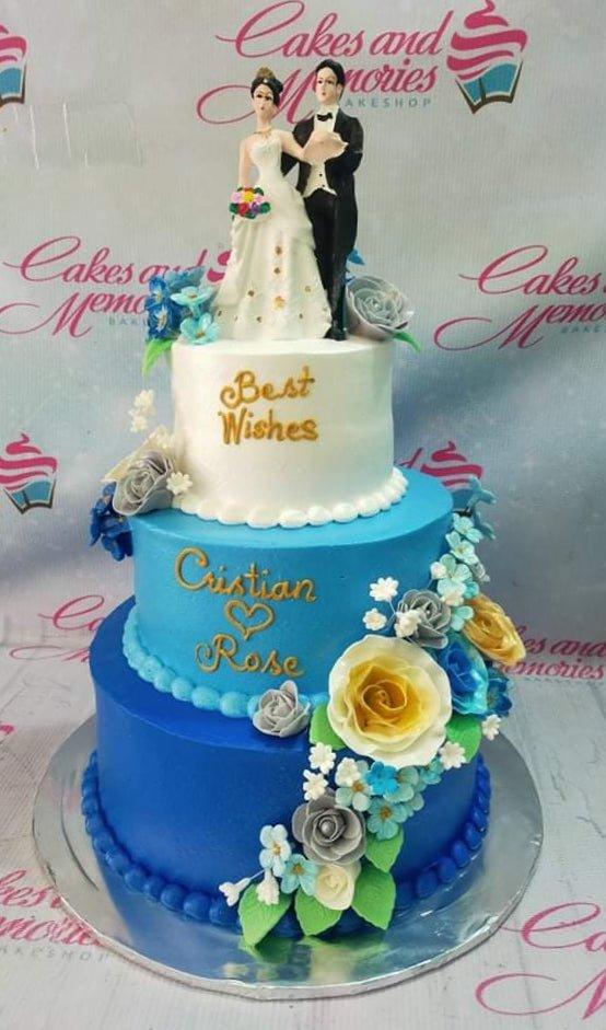 Elegant 3-tier wedding cake in blue and white gradient with a bride and groom topper and cascading sugar flowers.