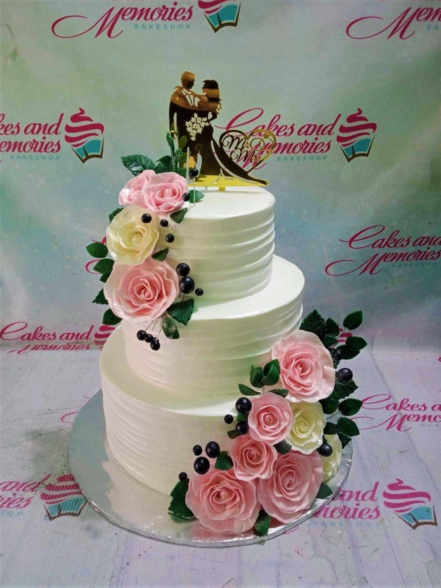 Elegant 3-tier white wedding cake with pink and cream gumpaste roses and a gold silhouette couple topper.