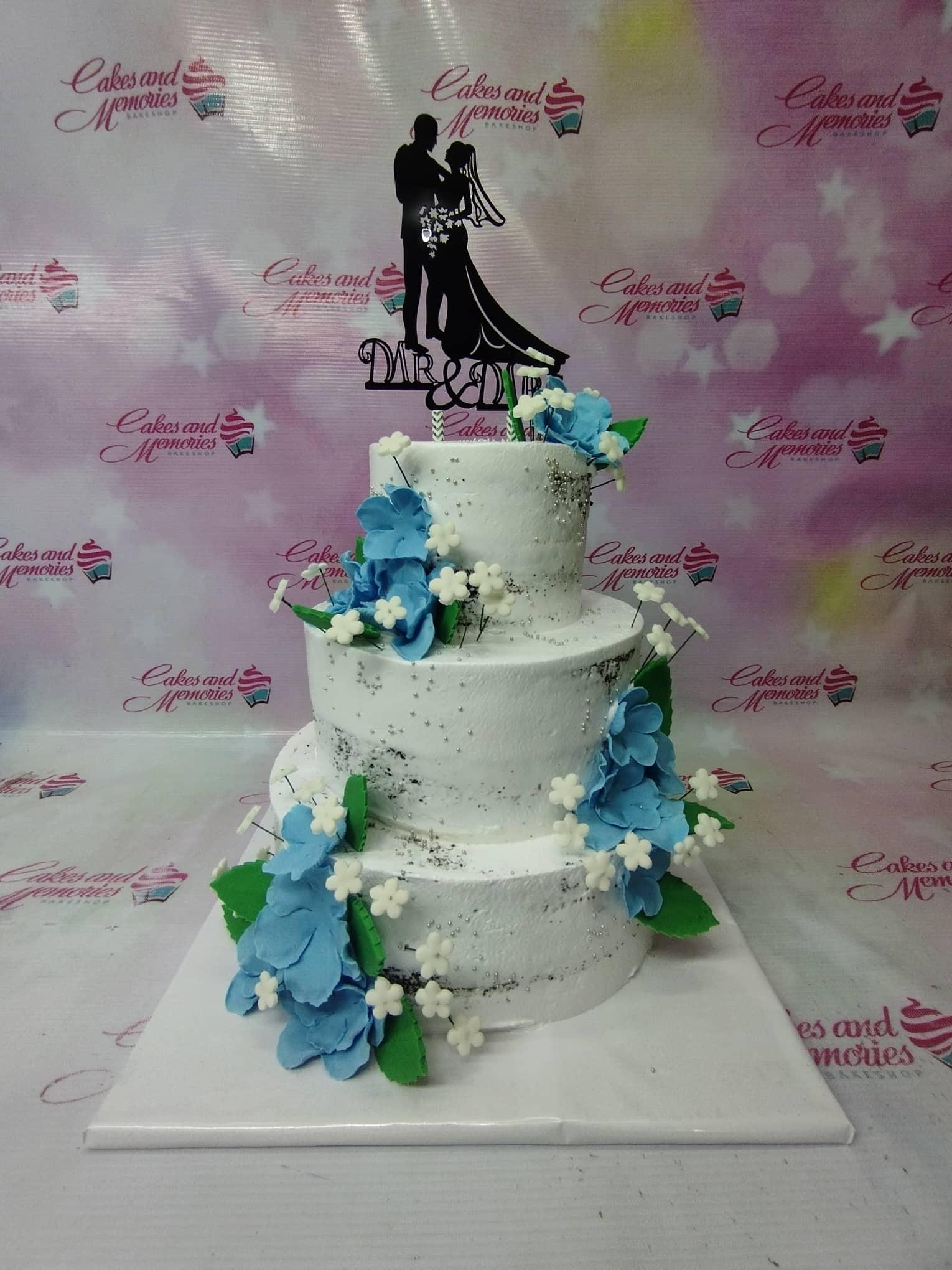 Three-tier white wedding cake with blue and white gumpaste flowers, silver dragees, and a black silhouette couple topper.