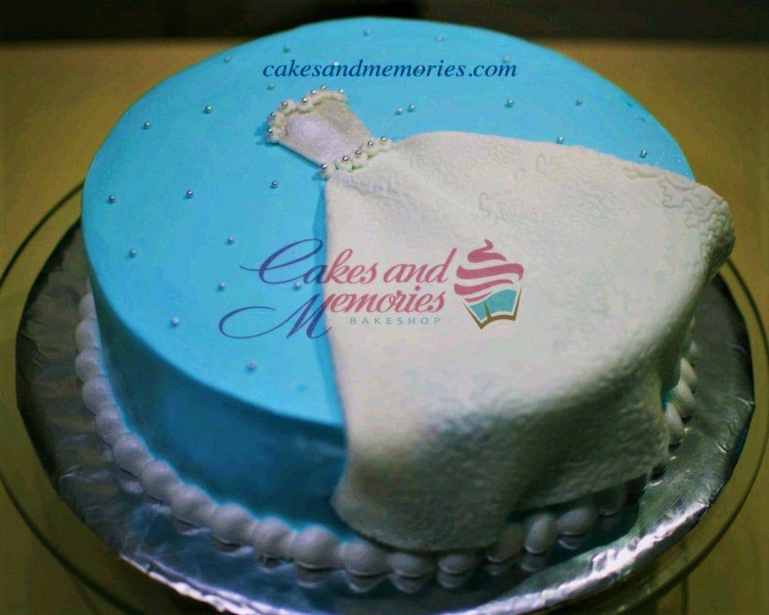 Light blue round cake featuring a white fondant wedding dress design with pearl accents and a white pearl border.