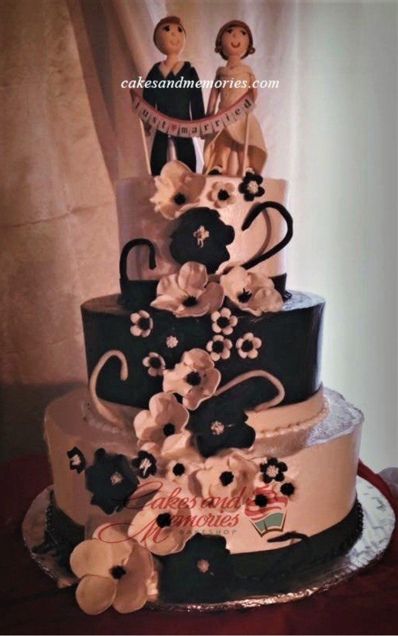 Elegant three-tier wedding cake with black and white floral decorations and a bride and groom topper holding a Just Married banner.