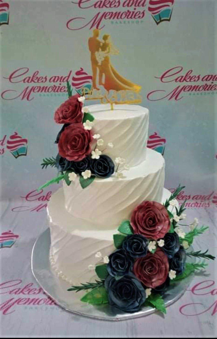 Elegant 3-tier white wedding cake with textured diagonal icing, dark red and navy blue roses, and a gold bride and groom topper.