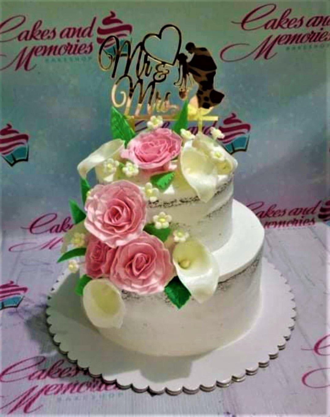 Two-tier semi-naked wedding cake with pink roses, white calla lilies, and a gold Mr & Mrs topper.