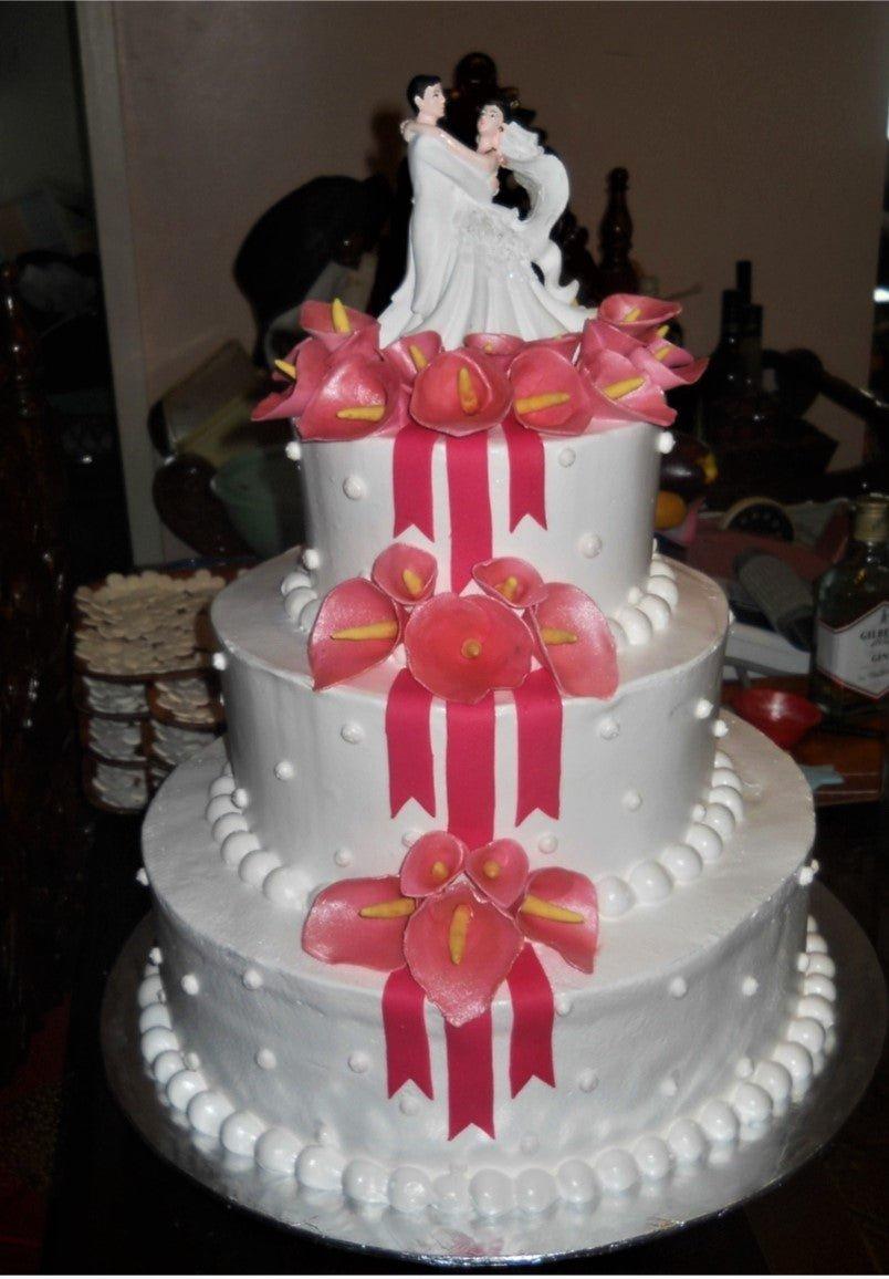 Elegant 3-tier white wedding cake with pink calla lilies, red ribbons, and a bride and groom topper.