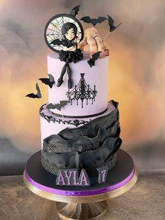 Two-tier lavender Wednesday Addams themed cake with black ruffles, bats, a chandelier silhouette, and a 3D Wednesday figure on top.