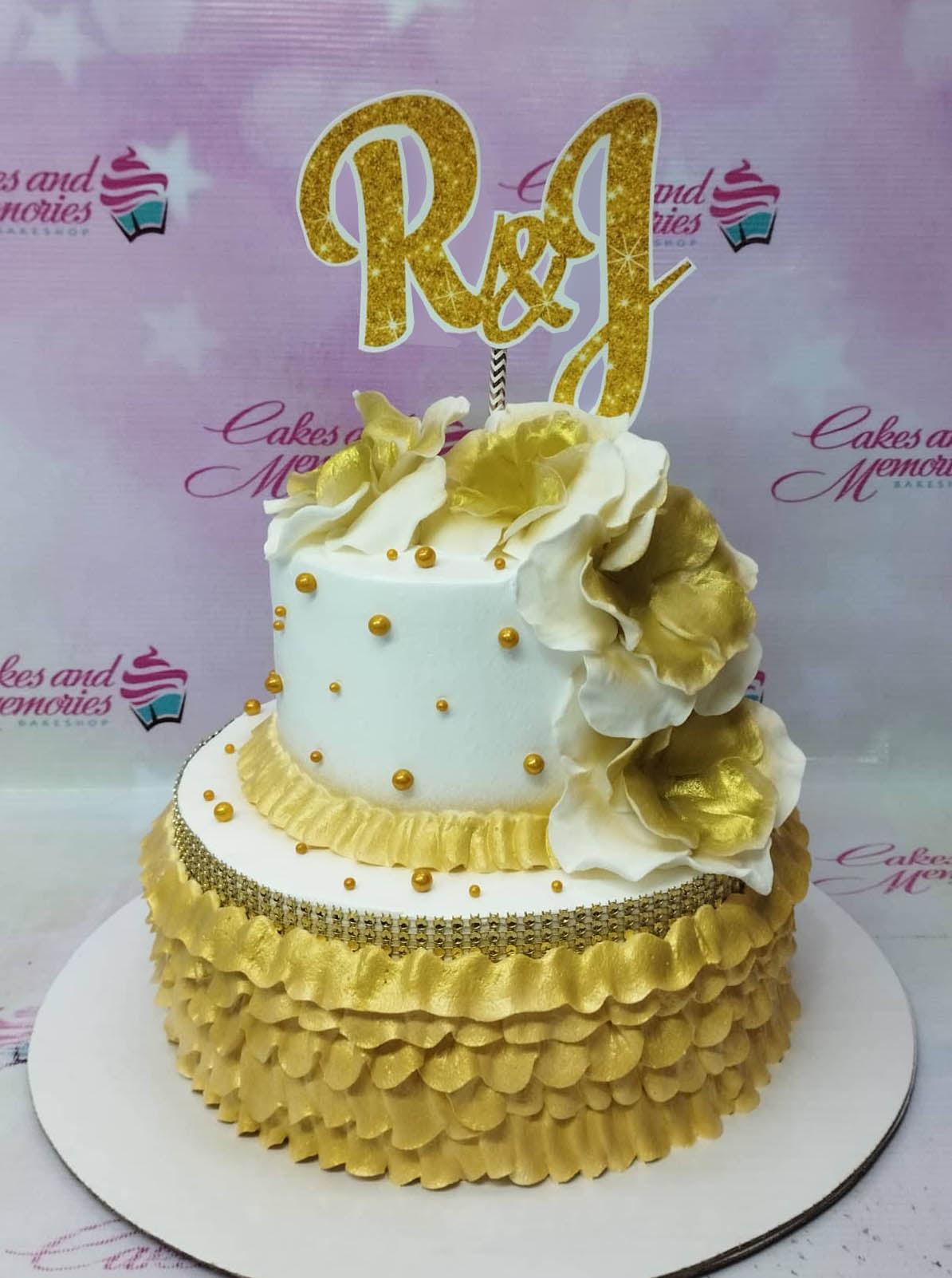 Elegant 2-tier white and gold anniversary cake with gold ruffles, large white and gold sugar flowers, and a glittery R&J topper.