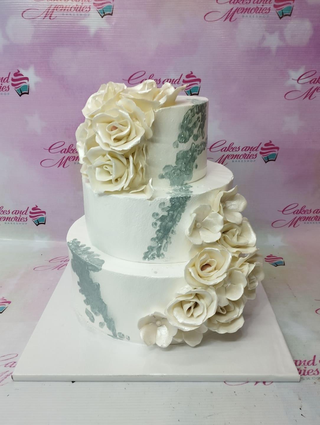 Elegant three-tier white wedding cake decorated with large white gumpaste roses and silver palette knife icing accents.