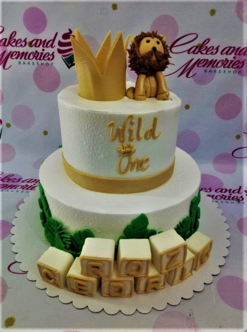White 2-tier Wild One lion birthday cake with a gold crown, fondant lion, green tropical leaves, and name blocks.
