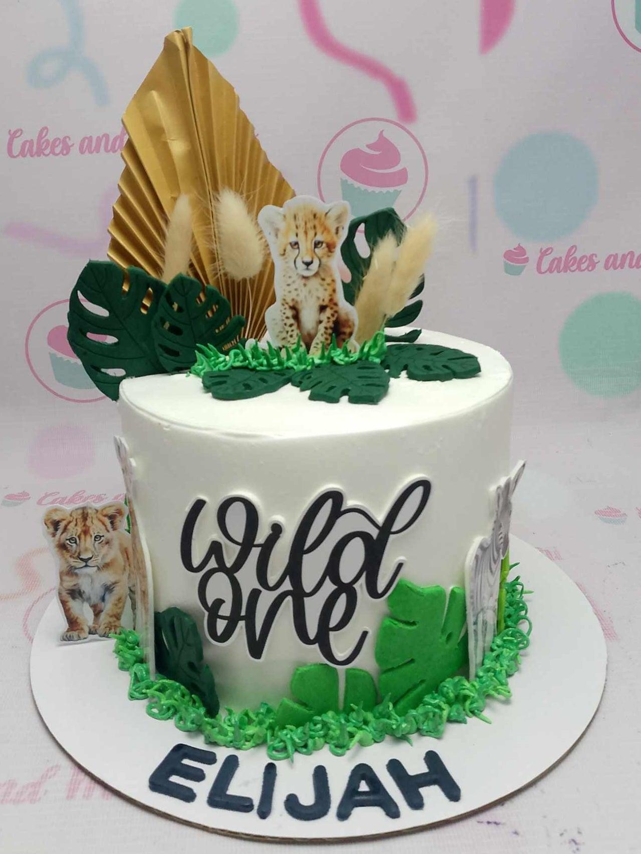 White 1-tier safari themed birthday cake with cheetah and lion printouts, green fondant leaves, and a gold fan topper.