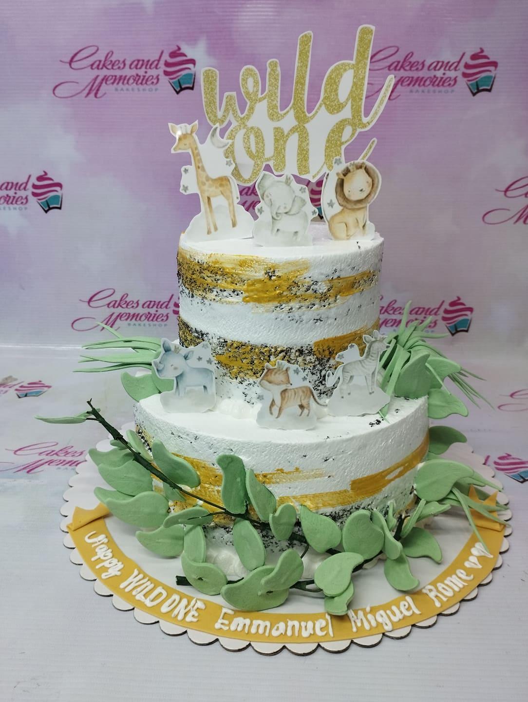 Two-tier white semi-naked cake with gold brushstrokes, green gumpaste leaves, and wild animal printout toppers for a first birthday.