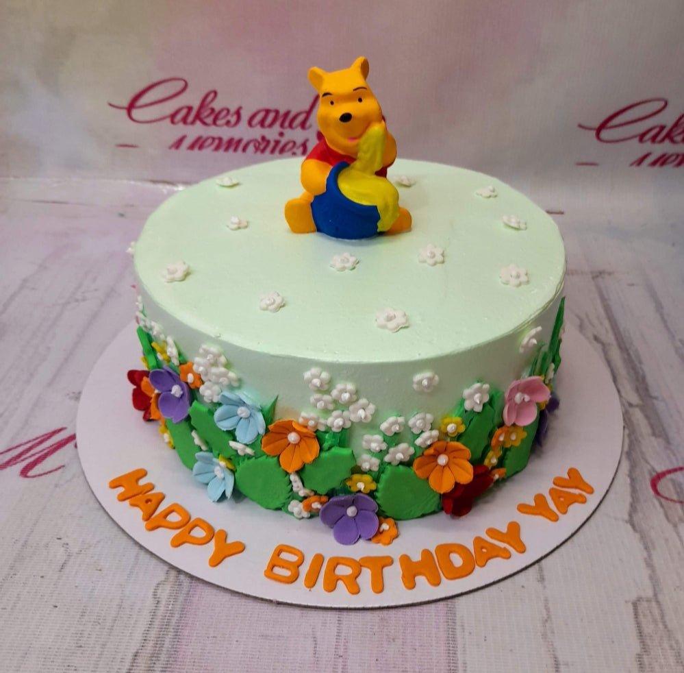 Winnie the Pooh birthday cake with a 3D fondant Pooh figure, colorful gumpaste flowers, and orange birthday message on a light green base.