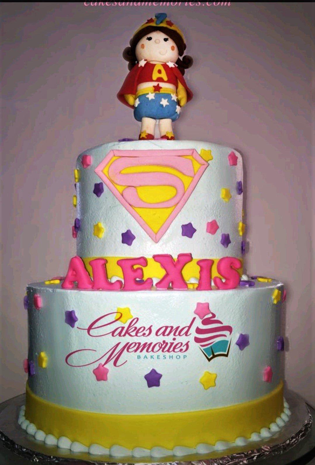 Two-tier light blue fondant cake with a Wonder Woman style girl topper, pink Superman logo, and colorful fondant stars.