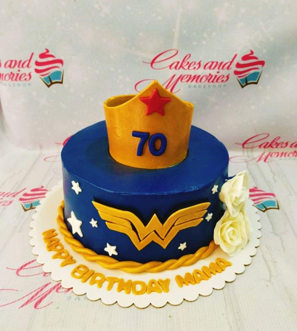 Wonder Woman themed birthday cake in navy blue with a gold crown topper, gold logo, and white roses for a 70th birthday.