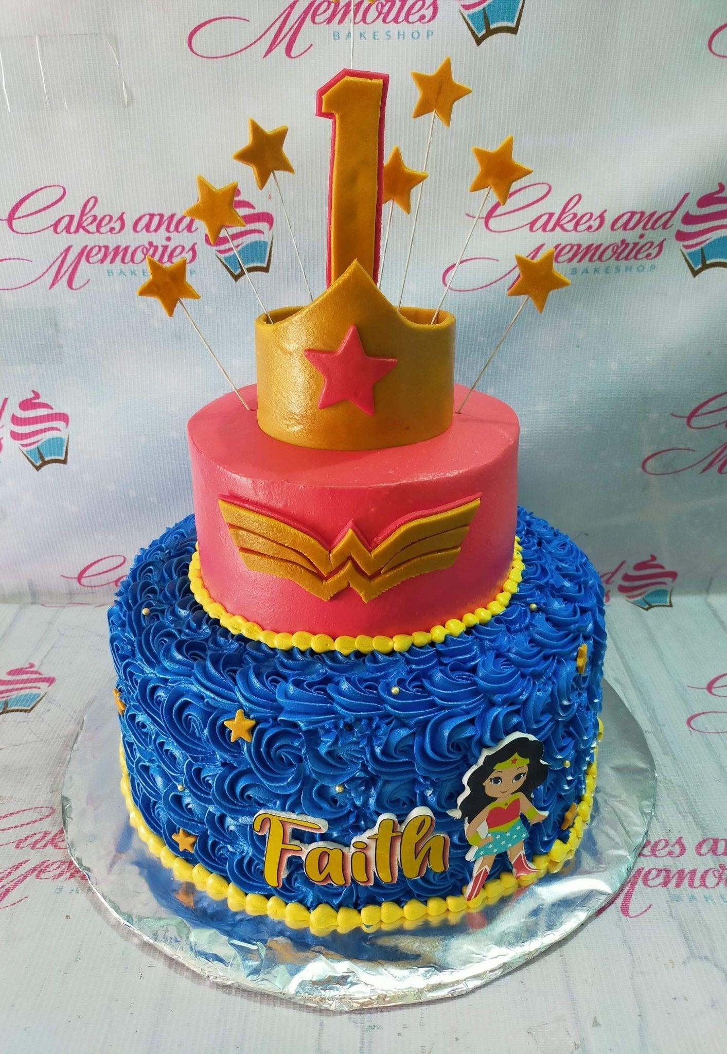 Two-tier Wonder Woman themed birthday cake with pink top tier, blue ruffled bottom tier, gold crown, and star toppers.