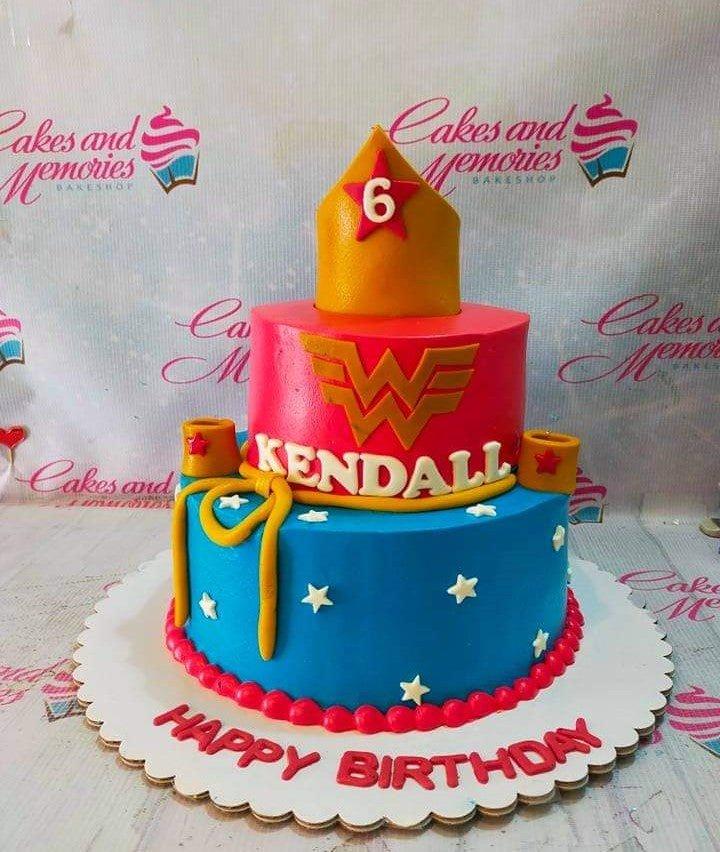 Two-tier Wonder Woman themed birthday cake with red and blue fondant, gold tiara topper, and lasso details.
