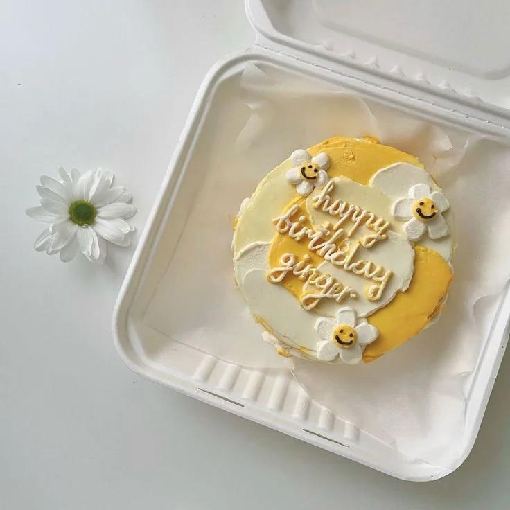 Yellow and white bento cake with piped daisy flowers and a happy birthday message in a white takeout box.