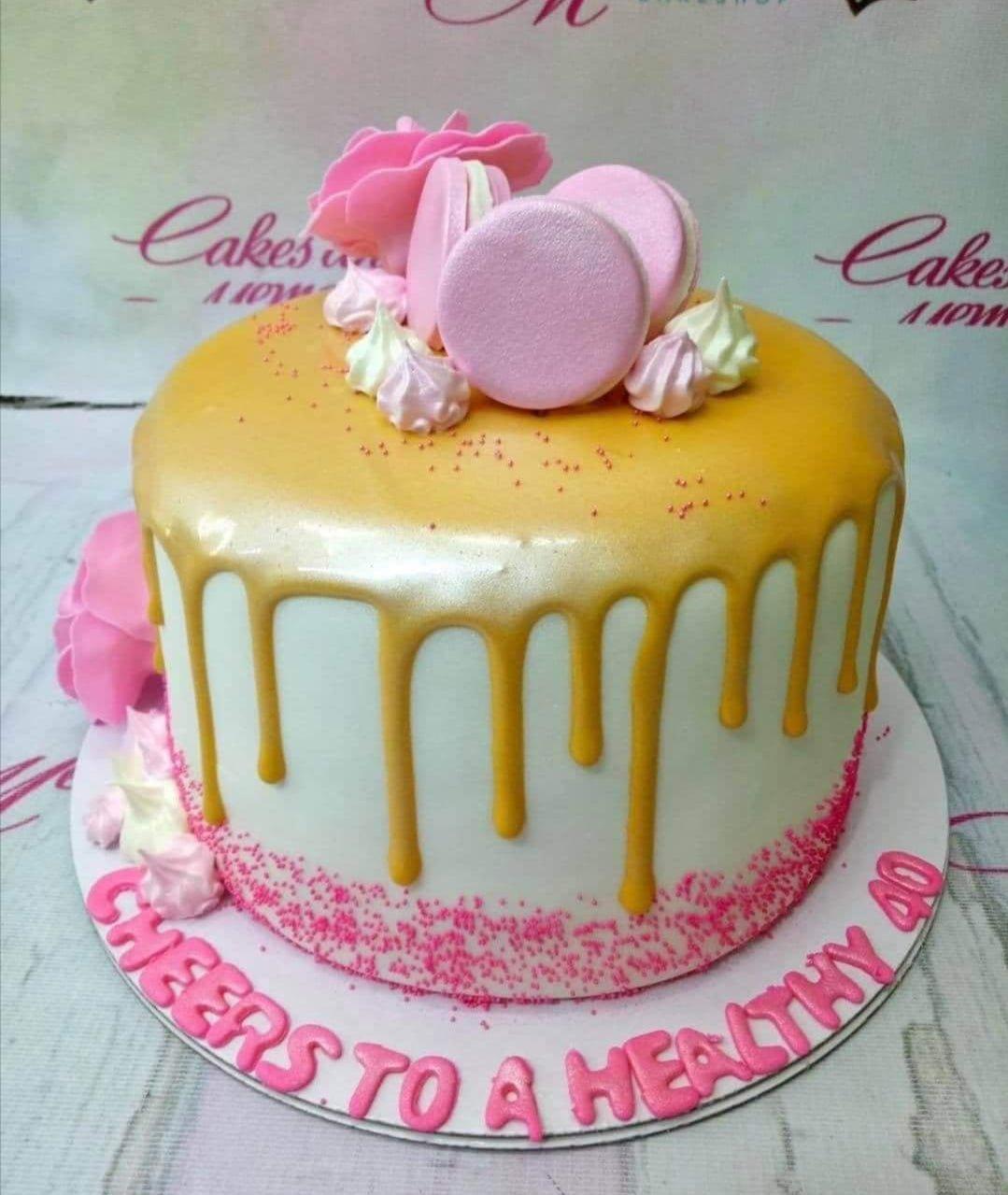 White 1-tier cake with yellow drip, pink macarons, meringue kisses, and pink gumpaste flowers with a birthday message on the base.