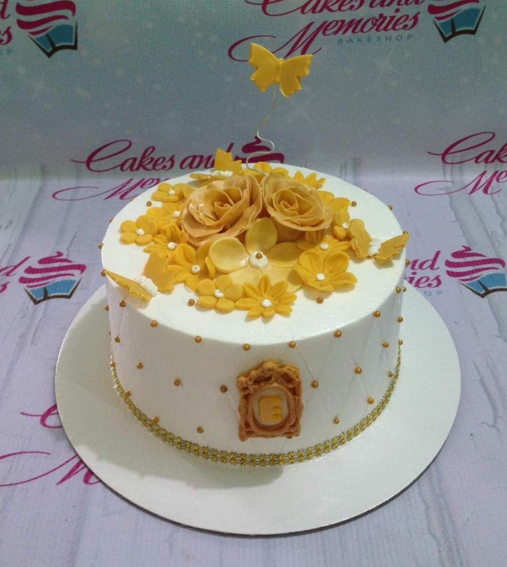 White round cake with yellow gumpaste roses, flowers, and butterflies, featuring a quilted pattern with gold dragees and a gold frame initial.