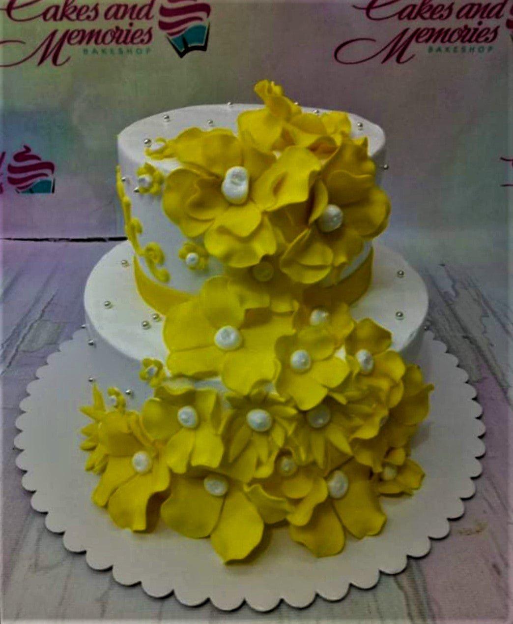 Two-tier white cake decorated with a cascading trail of large yellow gumpaste flowers and silver dragees.