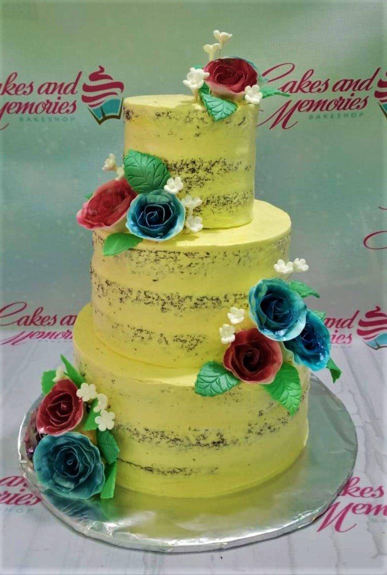 Three-tier yellow semi-naked cake decorated with red and teal gumpaste roses and green leaves.