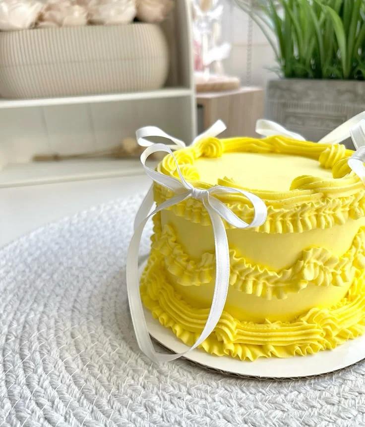 Yellow minimalist vintage cake with ruffled icing borders and white ribbon bows on a white textured surface.