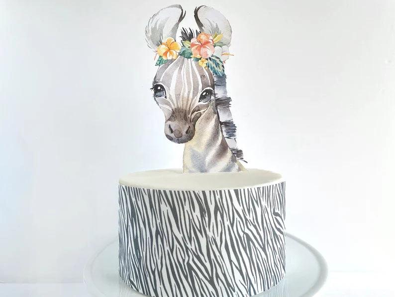White fondant cake with a zebra print side wrap and a watercolor zebra printout topper wearing a floral crown.