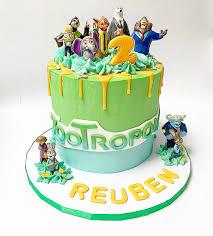 Zootopia themed birthday cake with green and blue icing, yellow drip, and various character figurines on top and around the base.