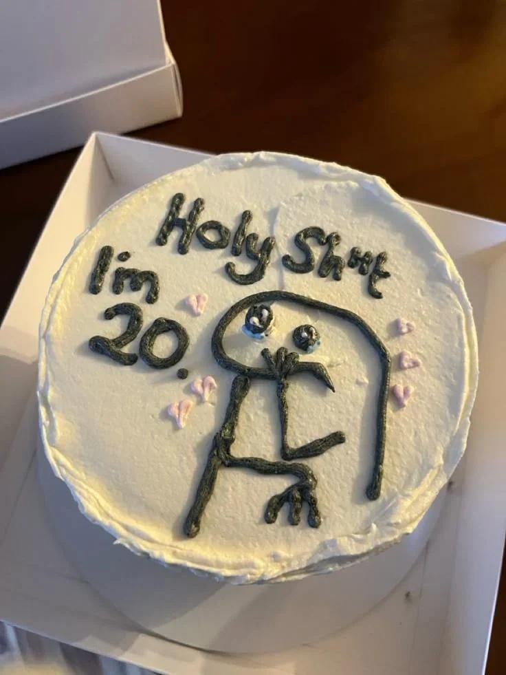 White bento cake with a black piped stick figure character and the message 'Holy Shit I'm 20' with small pink hearts.