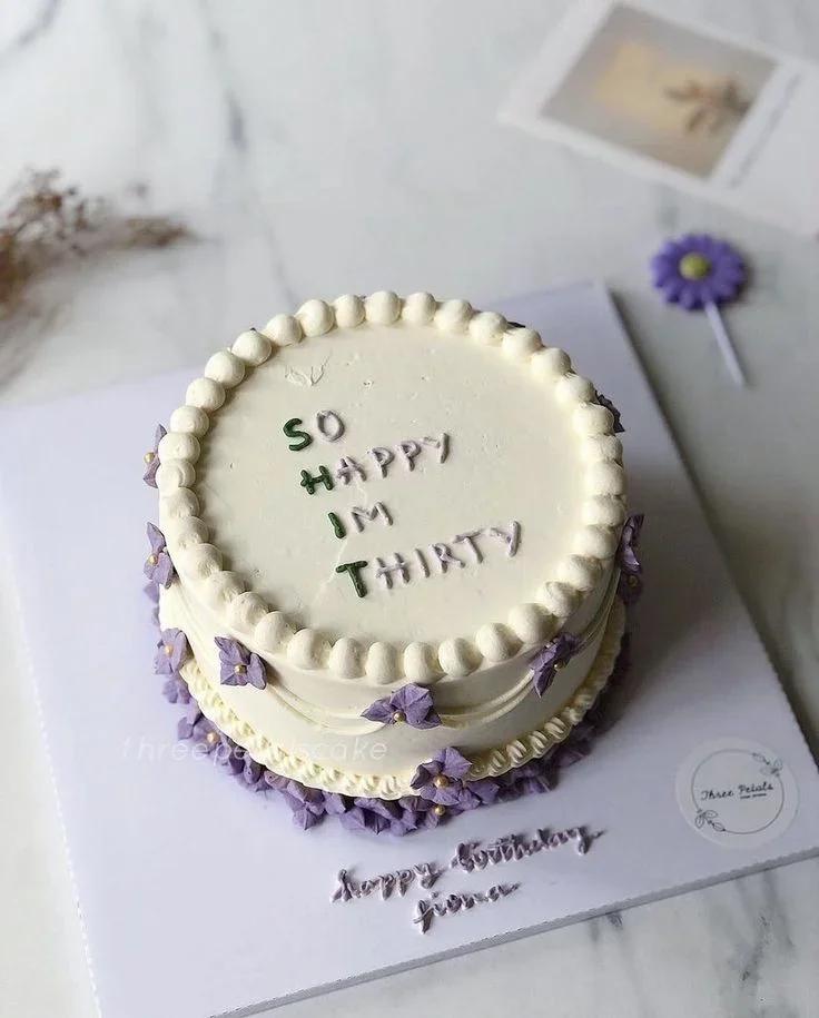 White minimalist birthday cake with purple icing flowers and a humorous message 'SO HAPPY IM THIRTY' in green and purple icing.