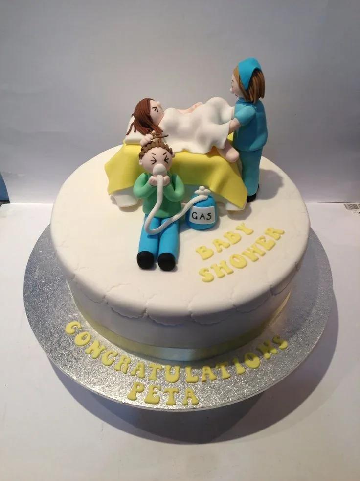 White fondant baby shower cake featuring 3D edible figures of a mother in labor, a father with a gas mask, and a nurse.