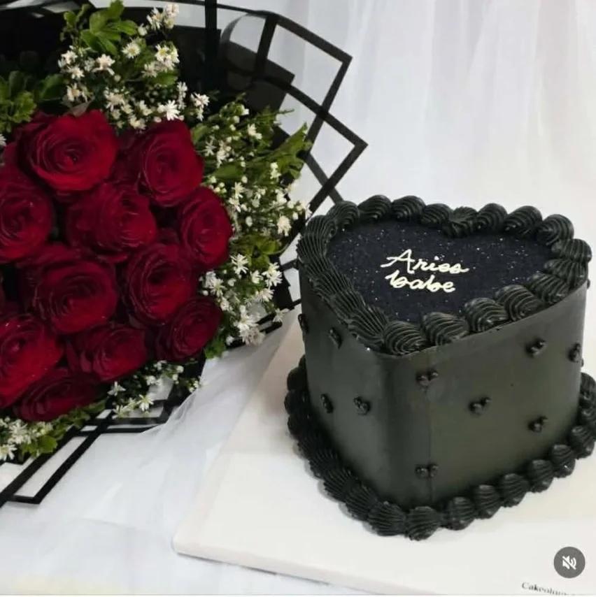 Heart-shaped black minimalist cake with white piped icing text and black rosettes, paired with a bouquet of red roses.