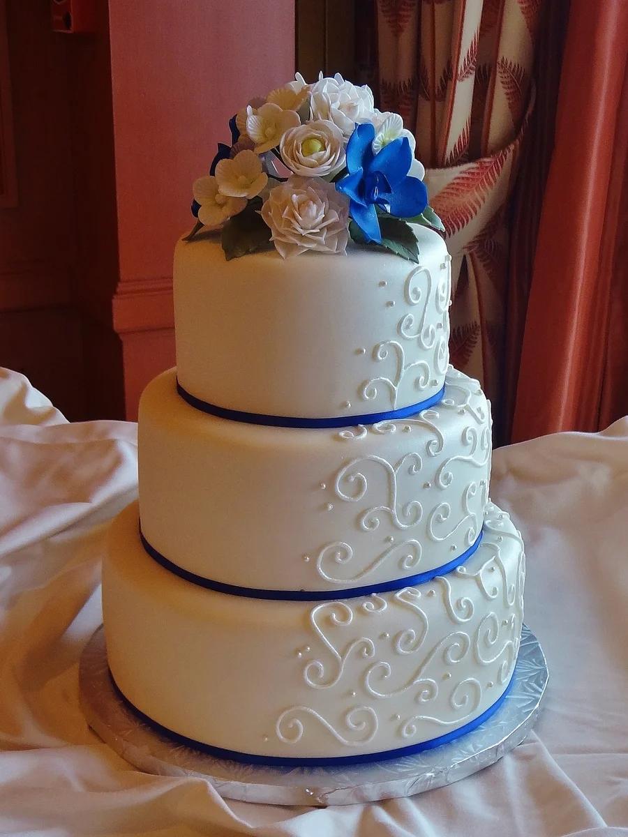 Elegant 3-tier white fondant wedding cake with blue and white sugar flowers and intricate white royal icing scrollwork.