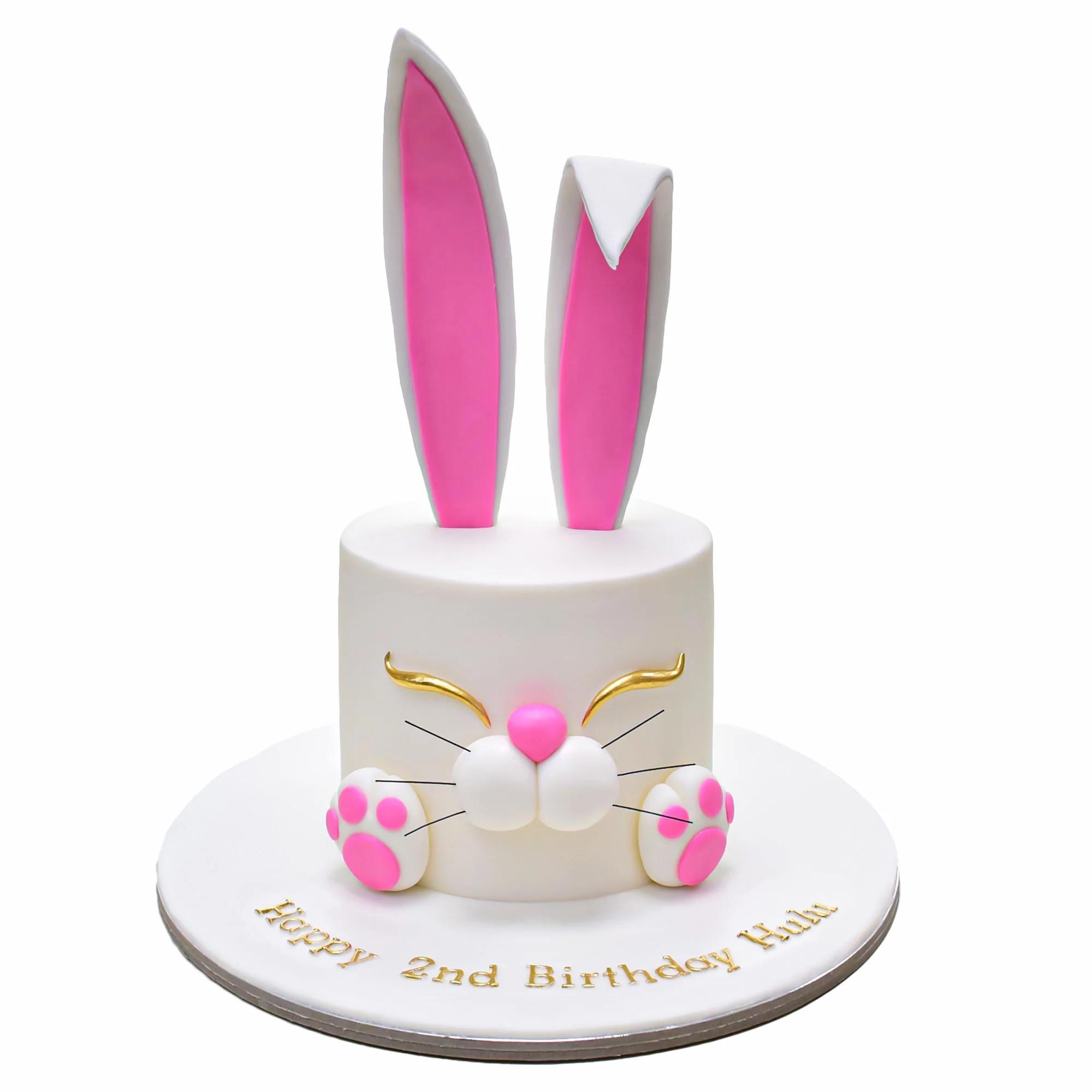 White fondant bunny cake with large pink and white ears, golden eyes, and pink paws on a white base board.