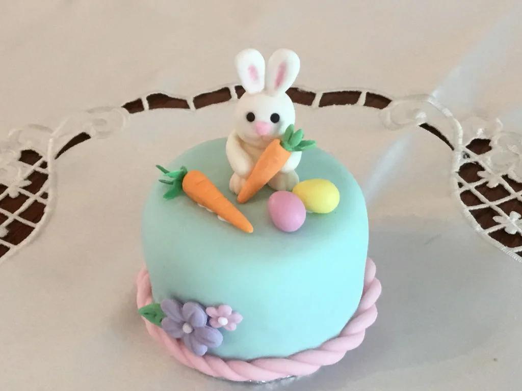 Small light blue fondant cake with a 3D bunny topper holding a carrot, decorated with fondant eggs, carrots, and flowers.
