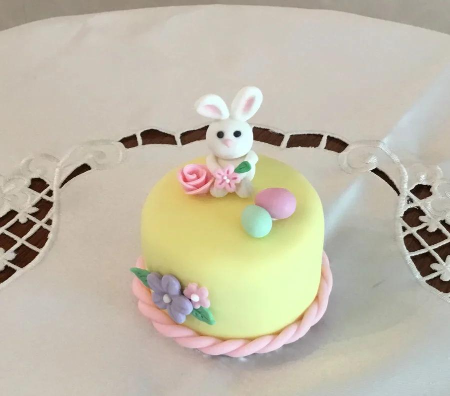 Small yellow fondant cake with a white bunny topper, pink and purple flowers, and pastel eggs.