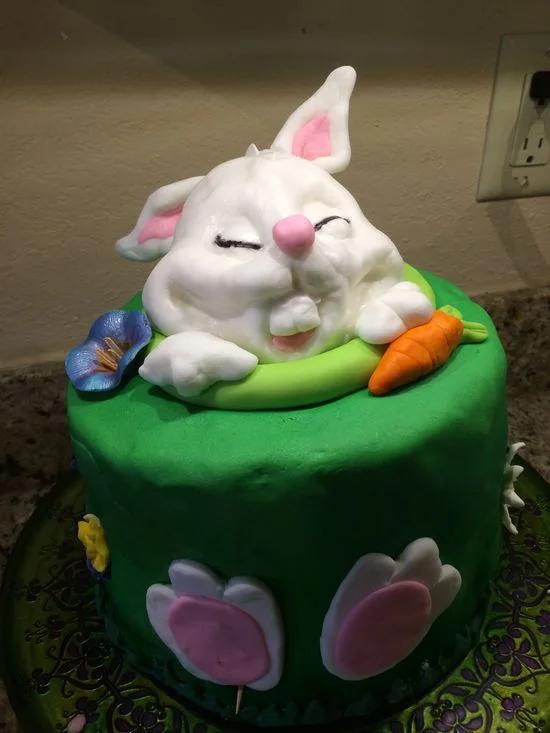Green fondant cake featuring a large 3D white bunny popping out of the top with a carrot and colorful flowers.