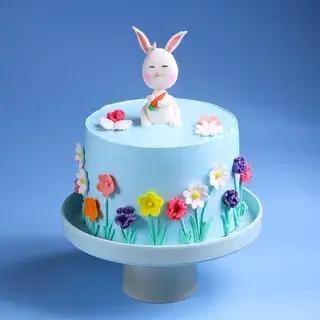 Light blue round cake decorated with colorful gumpaste flowers and a white bunny topper holding a carrot.